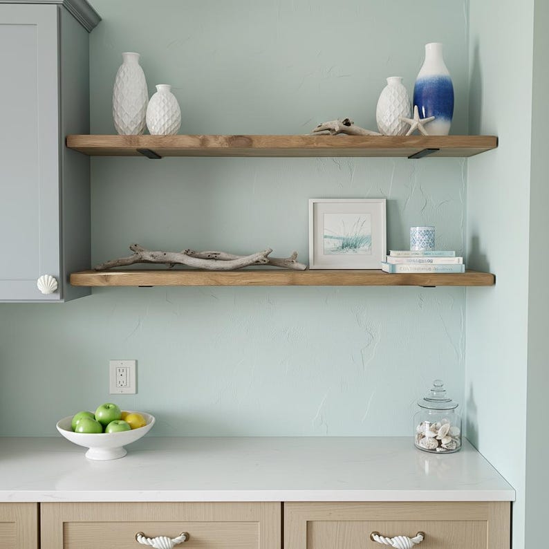 May include: A coastal-themed kitchen display featuring two wooden shelves with decorative items. The shelves hold white textured vases, a blue and white vase, a framed picture, books, and driftwood. A white bowl of green apples and lemons sits on the countertop.