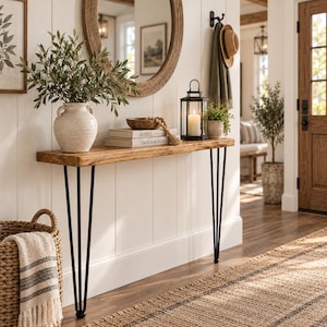 May include: A long, narrow console table with a natural wood top and four black metal hairpin legs. The table holds a white ceramic vase with greenery, a wooden bowl with beads, two stacked books labeled HOME, and a black metal lantern with a candle.