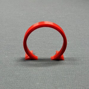 May include: A bright red, semi-circular object with a rounded top and two angular feet. The object is made of a solid material and stands upright against a gray background. The shape resembles a partial ring or a stylized bracket.