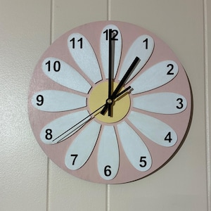 May include: A daisy-shaped wall clock with a light pink background and white petals. The clock face features black numbers and hands, with a yellow center. The clock is designed to resemble a daisy flower.