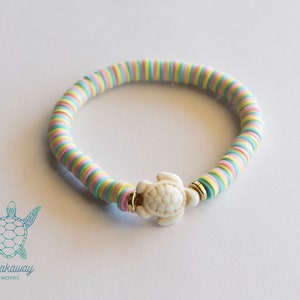 Peut inclure: Un bracelet coloré composé de perles rondes empilées en rose, jaune et bleu. Le bracelet a un charme de tortue blanche avec des accents dorés.