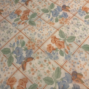 Könnte beinhalten: Weißer Stoff mit einem sich wiederholenden Muster aus blauen und orangefarbenen Blumen, grünen Blättern und Schmetterlingen. Das Muster ist auf einem Gitter aus braunen Linien angeordnet.