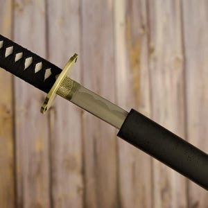 May include: A katana sword with a black handle wrapped with white diamond-shaped accents. The blade is silver, and the guard is gold. The sword is partially sheathed in a black scabbard, set against a wooden background.