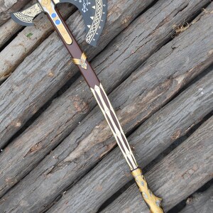 May include: A detailed, decorative axe with a dark gray blade featuring gold accents and intricate patterns. The handle is brown with gold and white detailing, and the axe rests on a wooden surface. The overall design suggests a fantasy or historical theme.