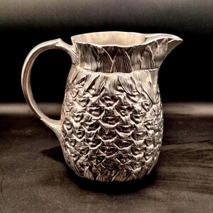 May include: A silver-colored pitcher with a pineapple design. The pitcher has a handle and a spout for pouring. The body of the pitcher is textured to resemble a pineapple, with a detailed leaf design at the top.