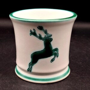 May include: A white ceramic cup featuring a dark green hand-painted illustration of a leaping deer on the front. The cup has a flared rim and a circular base, both accented with a solid dark green painted band around the edges of the ceramic.