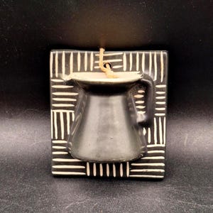May include: A black ceramic wall-mounted planter featuring a square backplate with a white etched dash pattern. A small pitcher-shaped vessel is attached to the center, and a tan twine loop is threaded through a hole at the top for hanging.