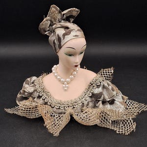 May include: A porcelain bust of a woman wearing a brown and white patterned turban, a pearl necklace, and a gold and brown lace collar. The bust is on a black background.