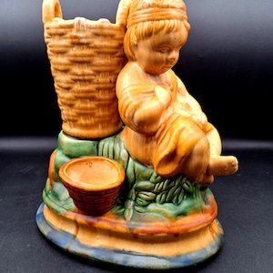 May include: A ceramic figurine featuring a child sitting beside a tall, woven-textured basket and a smaller, separate bowl-shaped basket. The piece is finished in shades of orange, green, and blue, with a sculpted base and detailed textures.