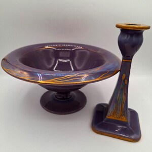 May include: A purple ceramic bowl with gold accents and a matching purple ceramic candle holder with gold accents. The bowl has a pedestal base and the candle holder has a square base.