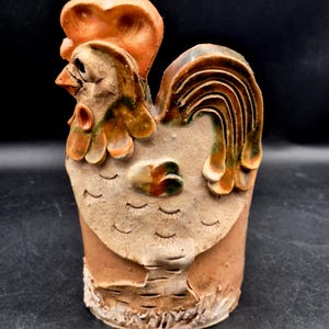May include: A cylindrical ceramic sculpture of a rooster with a light tan body and a textured base. The rooster features a prominent orange comb, a brown and green glazed tail, and incised feather details on the wings and body surface.