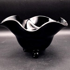 May include: A glossy black decorative bowl with a ruffled rim and three small, rounded feet. The bowl has a smooth, reflective surface, and the dark color gives it a sophisticated look. The bowl is likely made of glass or ceramic.