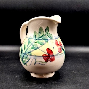 May include: Cream-colored ceramic pitcher with a rounded body and a handle. The pitcher is decorated with hand-painted red flowers, green leaves, and blue and green curved lines. The background is a pale yellow.