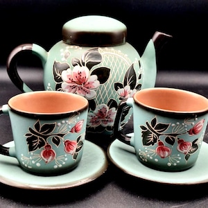 May include: A teal ceramic tea set featuring a teapot and two teacups with saucers. The set is decorated with pink floral designs and black accents. The interior of the cups is a warm brown color. The set is designed for serving tea.