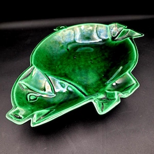 May include: A glossy, emerald green ceramic dish in the shape of a fish. The dish has a deep, open center and detailed features, including fins and a defined face. The ceramic piece is set against a black background.
