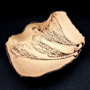 May include: A beige, free-form ceramic bowl with a black, organic design. The bowl has a dark brown rim and a textured surface. The design resembles a stylized plant or fern. The bowl is on a black background.