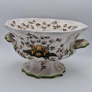 May include: A white ceramic bowl with green and yellow floral designs. The bowl has two handles and a pedestal base.