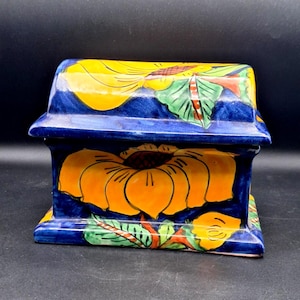 May include: A ceramic box with a hinged lid, featuring a vibrant floral design. The box is primarily blue, with large yellow sunflowers and green leaves. The design is hand-painted, showcasing a detailed and colorful aesthetic.
