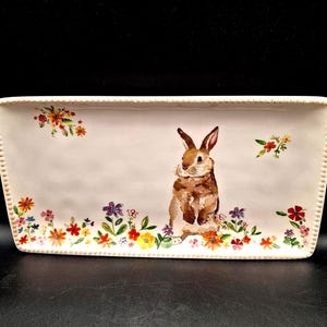 May include: Rectangular ceramic serving platter with a painted bunny and floral design. The bunny is brown and white, surrounded by colorful flowers. The platter has a beaded edge and is perfect for Easter.