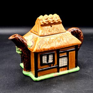 May include: A ceramic teapot shaped like a cottage with a thatched roof, brown walls, and black trim. The spout and handle resemble textured tree trunks, and the base is painted light green. The lid forms the roof of the house structure.