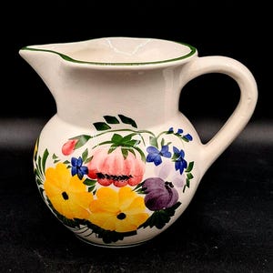 May include: A white ceramic pitcher with a green rim and a floral design. The flowers are yellow, pink, purple, and blue.
