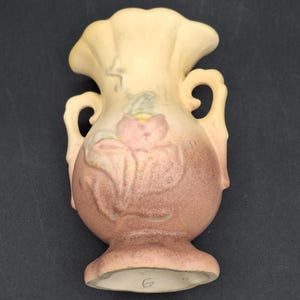 May include: A small, cream-colored ceramic vase with two handles. The vase has a floral design with a pink flower and green leaves. The vase has a brown glaze and is marked with a "G" on the bottom.
