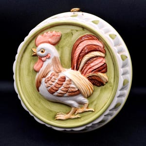May include: A round ceramic wall plaque featuring a raised, three-dimensional rooster in white, brown, and orange tones. The rooster is set against a light green background, surrounded by a white scalloped border with green leaf accents and a gold hanger.