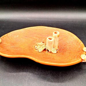 May include: A brown ceramic tray with two small, white ceramic cylinders in the center. The tray has two gold handles on the sides and a gold leaf design on the front.