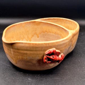 May include: A tan ceramic serving bowl with a divided interior and a glossy finish. The exterior features a sculpted, three-dimensional red lobster accent attached to the side. The glaze has a dripped, variegated appearance in shades of light brown.