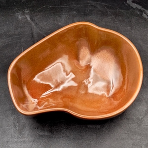 May include: A small, irregular, organic-shaped ceramic bowl with a glossy, warm amber-brown glaze. The interior surface features smooth, undulating curves and reflective highlights, showcasing a deep, rich color gradient throughout the piece.