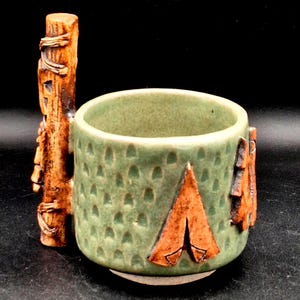 May include: A handmade ceramic mug with a green exterior and a brown tent design. The mug has a textured surface with small, evenly spaced indentations. A brown, carved wooden handle is attached to the side.