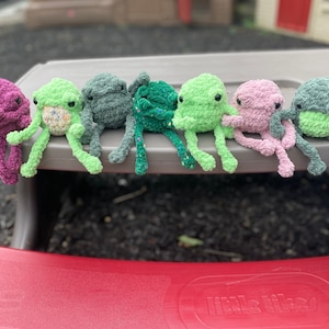 May include: Seven crocheted frog plushies in various shades of green, pink, and purple. The plushies are sitting on a gray table with their legs dangling over the edge.