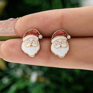May include: A pair of gold-toned Santa Claus stud earrings. Each earring features a white face with a gold outline, a red glitter hat, and a small clear gem. The earrings are designed for pierced ears and are suitable for the holiday season.