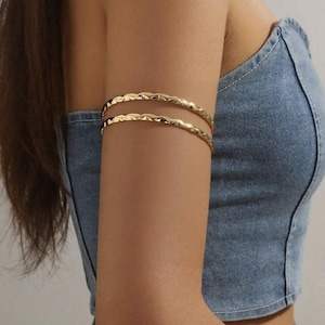 Puede incluir: Dos brazaletes dorados con un diseño ondulado. Los brazaletes se llevan en la parte superior del brazo. La modelo lleva un top de mezclilla.