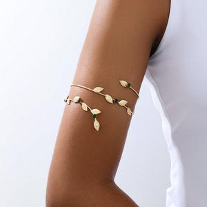 May include: Gold-toned arm cuff featuring a delicate leaf design with small green accents. The arm cuff is worn on the upper arm and has a minimalist, nature-inspired aesthetic. The jewelry is shown on a person's arm.