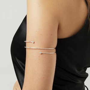 May include: A silver arm cuff with two delicate bands. The upper band has a pointed end, and the lower band features a small, round, clear gem. The jewelry is worn on the upper arm.