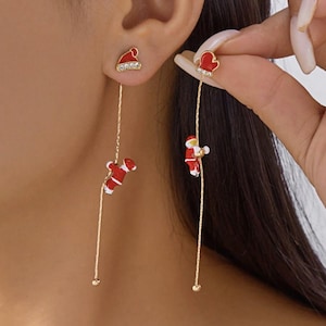 Puede incluir: Pendientes navideños dorados. Un pendiente presenta un perno de gorro de Papá Noel rojo con un Papá Noel colgante que sube por una cadena dorada. El otro pendiente tiene un perno de mitón rojo con un Papá Noel colgante. Los pendientes son festivos.