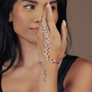 May include: A silver hand chain with a delicate leaf design. The chain extends from a ring on the middle finger, across the back of the hand, and connects to a bracelet. The leaves are adorned with small, sparkling stones.