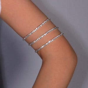 Può includere: Un bracciale da braccio argentato con tre file di scintillanti strass. Il bracciale è indossato sulla parte superiore del braccio e riflette la luce. Un accessorio glamour ed elegante, di circa 15 cm di diametro.