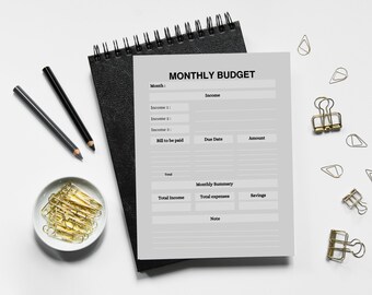 May include: A gray and white printable monthly budget planner with sections for income, bills to be paid, and a monthly summary. The planner is on a black notebook with a spiral binding.