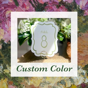 May include: A white table number card with a scalloped edge and the word "table" and the number "8" in green. The card is held by a gold-colored metal holder. The text "Custom Color" is below the card.