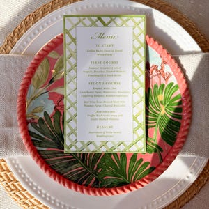 Puede incluir: Una mesa puesta con un plato blanco, un plato coral con un diseño de hojas tropicales y una tarjeta de menú. La tarjeta de menú tiene un borde de enrejado verde y enumera una comida de varios platos. Los platos están sobre un mantel individual tejido.