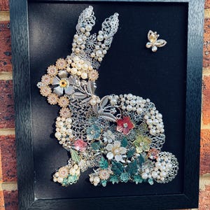 May include: A framed artwork depicting a rabbit silhouette made from vintage jewellery. The rabbit is embellished with pearls, rhinestones, and floral details in various colours. A small butterfly brooch is also included. The artwork is set against a black background within a black frame.