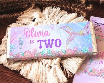 May include: A mermaid-themed chocolate bar wrapper with a purple and pink background. The wrapper features a mermaid tail, seashells, and the text "Olivia is Two".