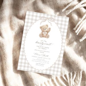 May include: A baby shower invitation with a teddy bear illustration. The invitation features a light gray gingham pattern and an oval frame with a bow. The text reads "We can bearly wait for baby Smith to arrive."