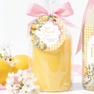 May include: A yellow candle in cellophane, tied with a pink ribbon and a decorative tag. The tag has a lemon and floral design with the words "Thank you". Two lemons and white flowers are in the foreground.