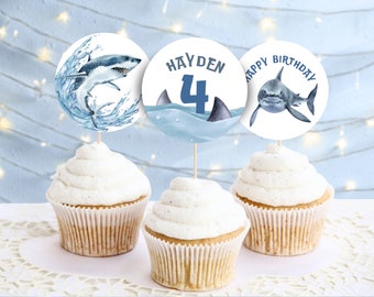 May include: Three blue and white cupcake toppers with a shark theme. One topper says "Happy Birthday", another says "Hayden 4", and the last one has a shark jumping out of the water.