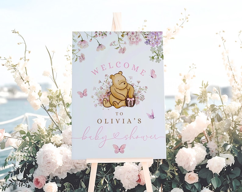 May include: A welcome sign for a baby shower with a Winnie the Pooh illustration. The sign is white with pink and purple floral accents and the text "Welcome to Olivia's Baby Shower".