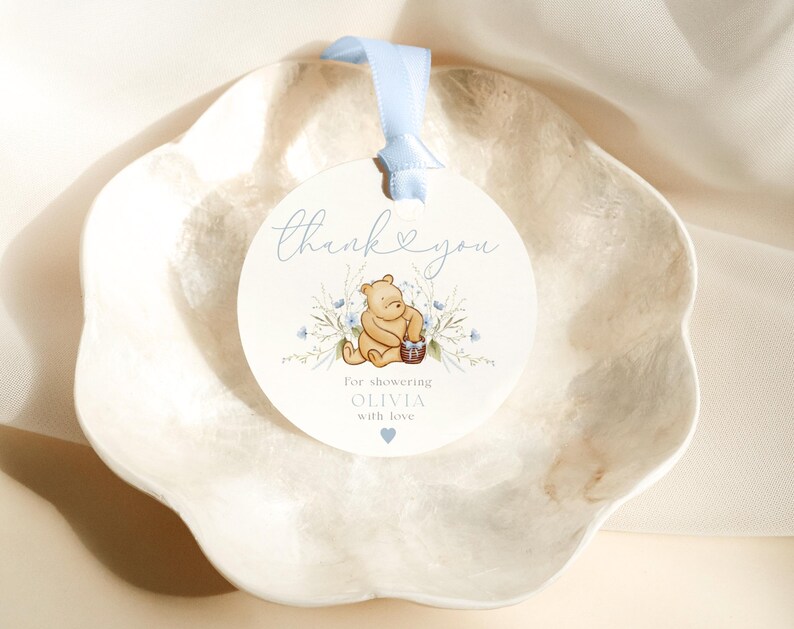 Pu&ograve; includere: Un'etichetta rotonda di ringraziamento con un nastro blu. L'etichetta presenta un orso Winnie the Pooh dei cartoni animati che tiene un cestino di miele. Il testo sull'etichetta dice "Grazie per aver festeggiato Olivia con amore".