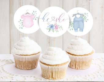 May include: Three cupcakes topped with white frosting, each adorned with a circular topper. The toppers feature watercolor illustrations of baby clothes, floral arrangements, and the text "he she". The color palette includes pink, blue, and green.